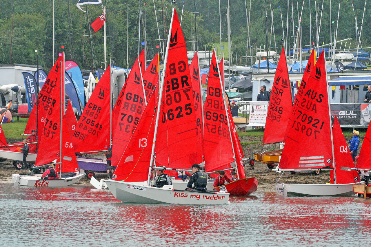 A fleet of Mirror dinghies lauching from a sandy beach with a sailing club in the background