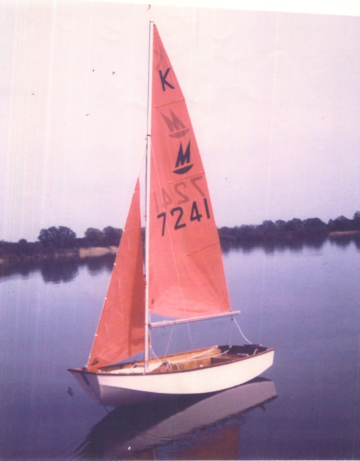 A white wooden Mirror dinghy with Bermuda rig with both sails hoisted, but no crew aboard on a long painter on a lake on a calm day