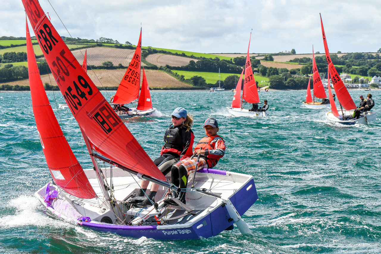 A purple & white Mirror dinghy being sailed to windward by two children on a very windy day viewed from the leeward side 