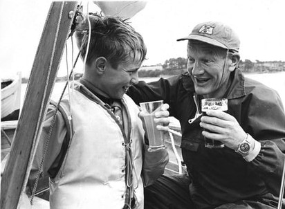 An adult man and his young crew with glasses of drink aboard their Mirror dinghy celebrating