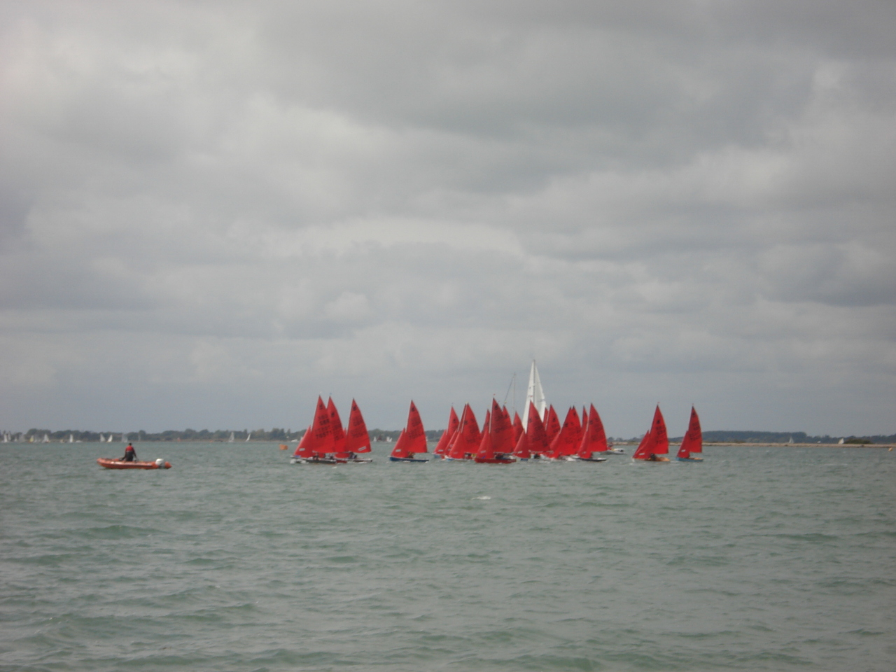 A fleet of Mirror dinghies starting a race in the distance under a cloudy sky