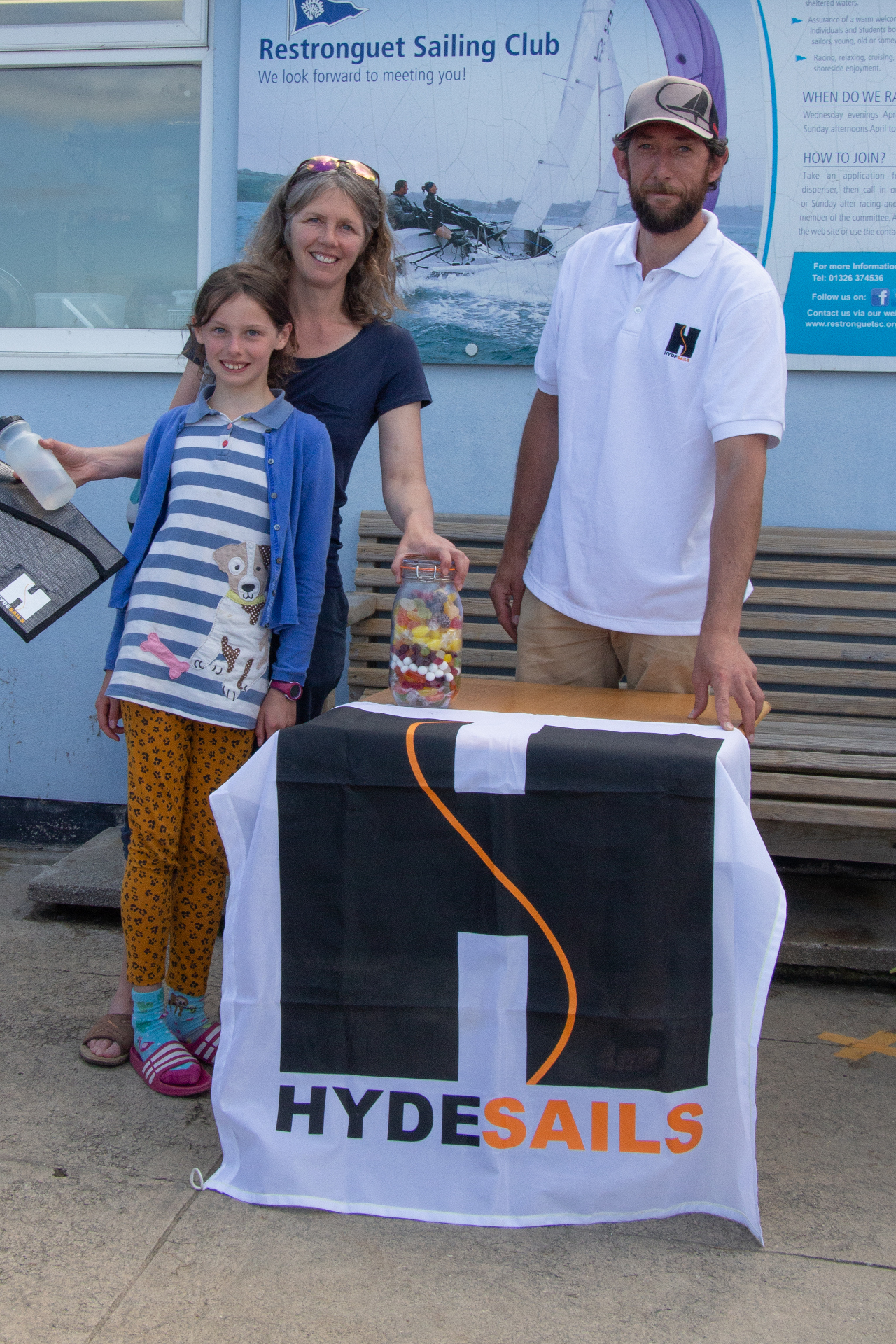 A young girl and her mum, receiving a voucher from a man with a large jar of sweets on a table with a Hyde Sails banner