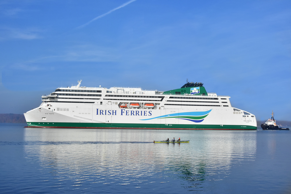 An ferry owned by Irish Ferries sailing on calm water with a tug close behind and a rowing skiff in the foreground