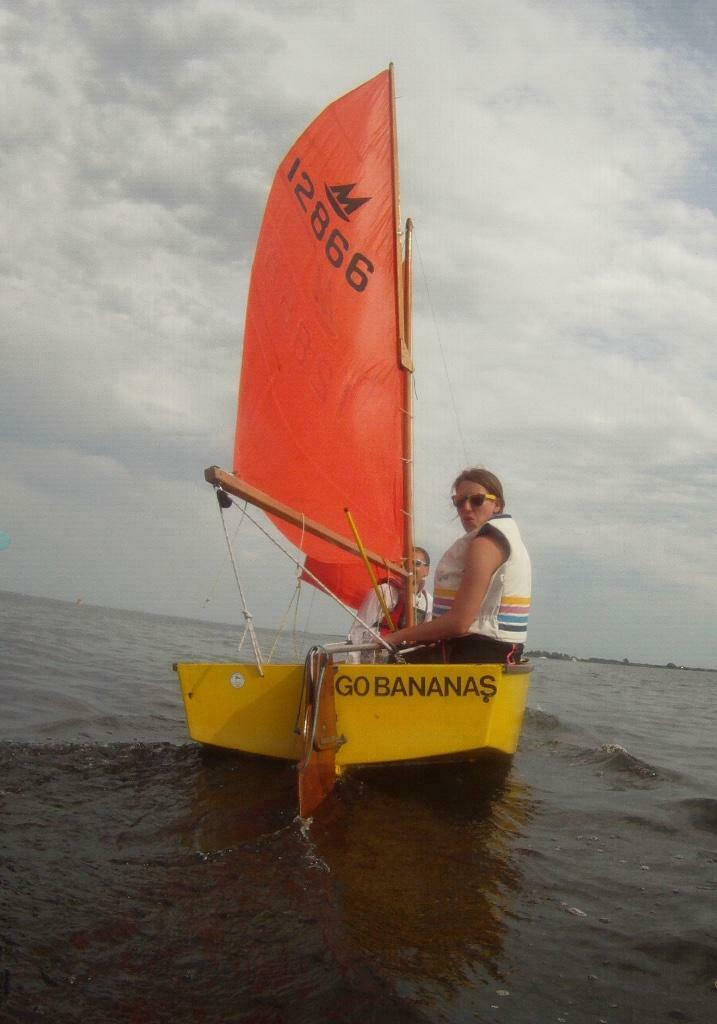 A yellow wooden Mirror dinghy sailing upwind, away from the camera, with helm looking over the stern towards the camera