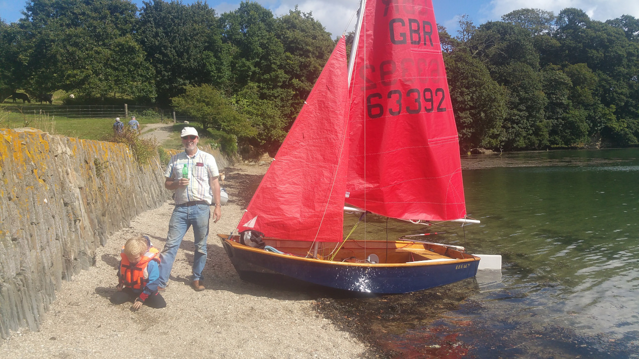 A very shiny blue wooden Mirror dinghy with sails hoisted gently resting its bow on a beach in the sunshine