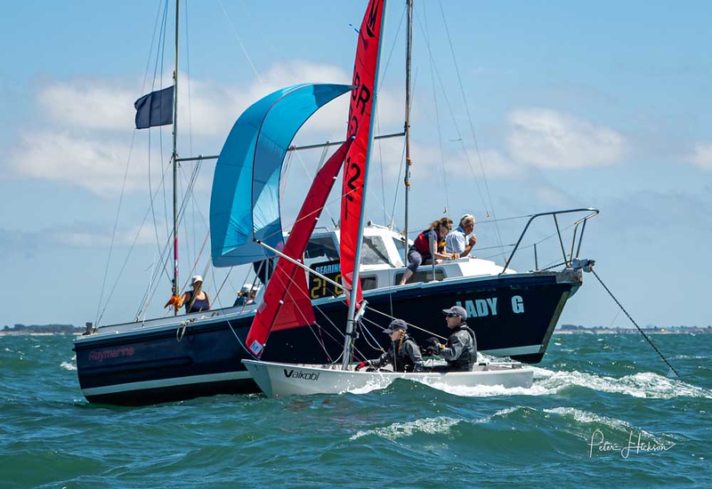 A white GRP Mirror dinghy crossing the finish line with spinnaker set and committee boat in the background